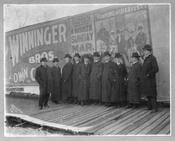 A group of men in long coats stands on a wooden platform in front of a large billboard.