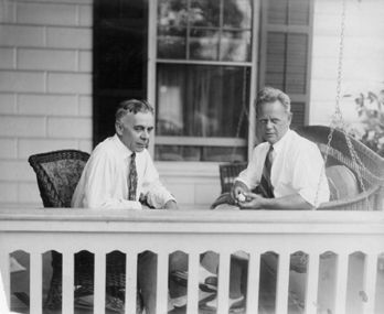 Two men in white shirts sit on a porch, one holding a cigar, with a window behind them.