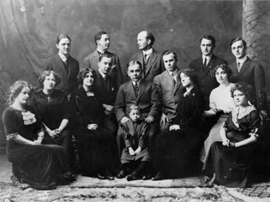 Formal group portrait of twelve people seated and standing in dark attire, posed against a studio backdrop