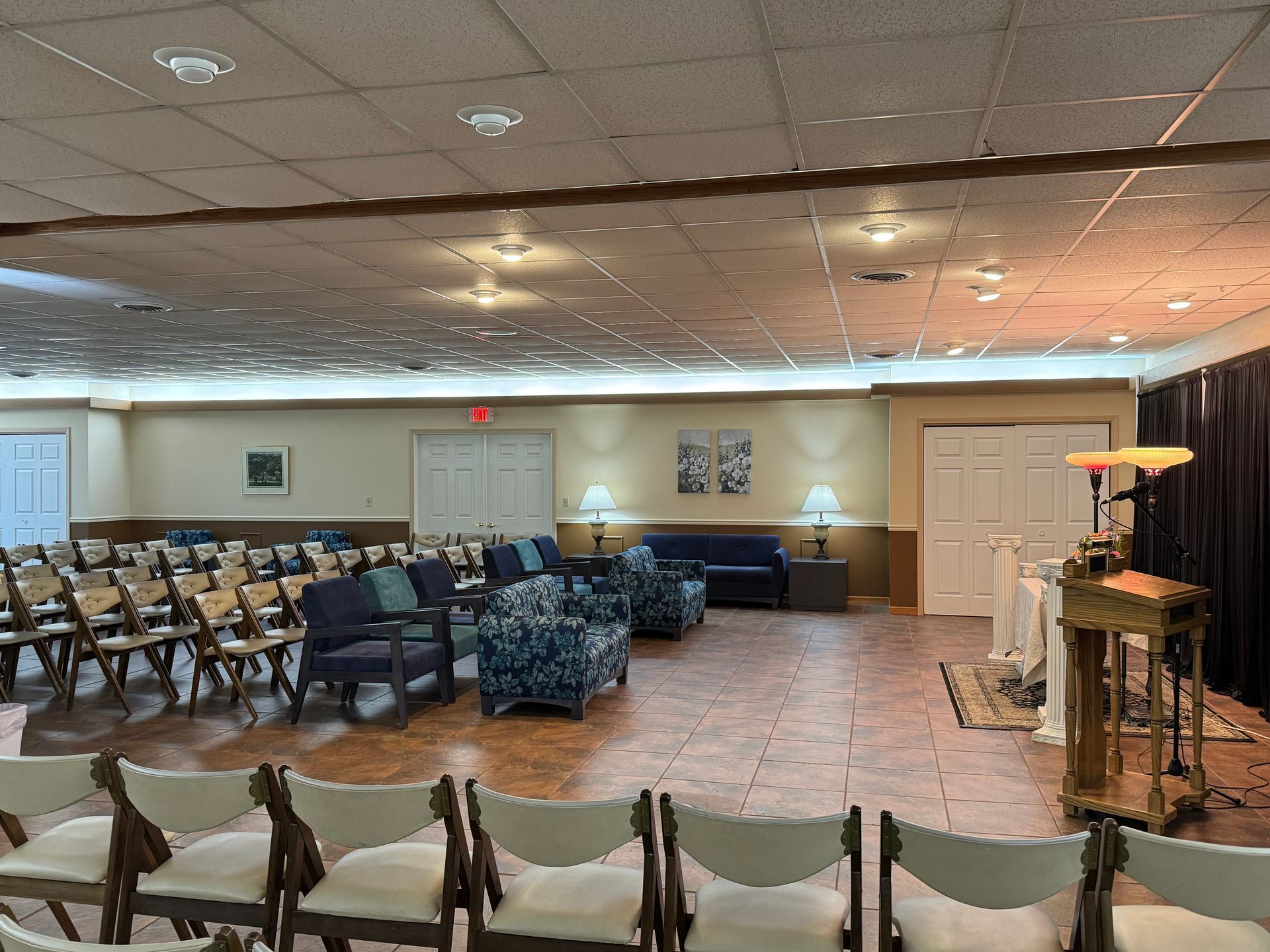 Empty community hall with rows of chairs, beige walls, tiled floor, and a podium at the right.