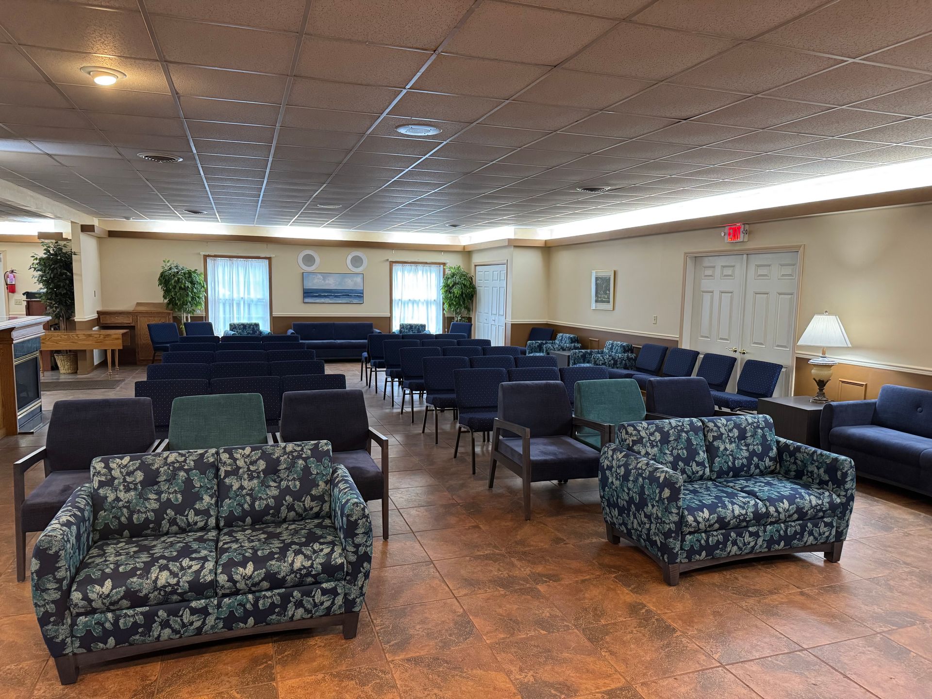 Empty community room with rows of chairs, patterned sofas, and a stage area with windows and lamps.