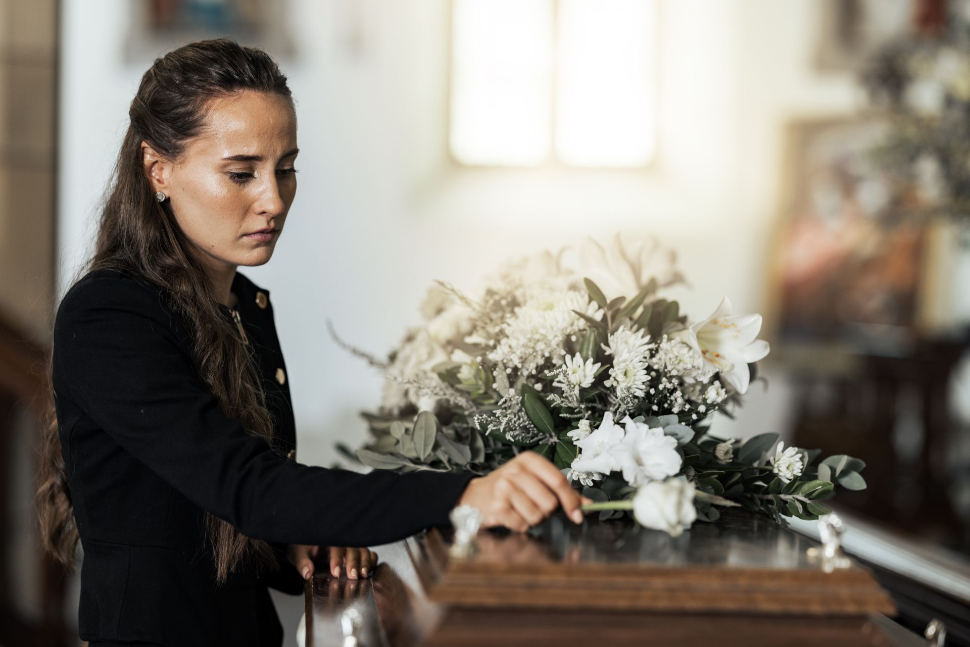 Woman placing white flowers on a wooden casket in a softly lit room