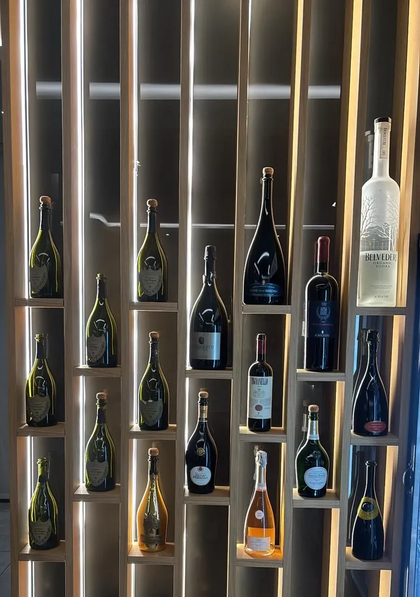 Una scaffalatura verticale in legno a griglia, piena di bottiglie di vino e champagne di vario tipo, illuminata da una retroilluminazione.