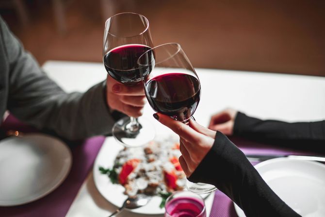 Due persone brindano con bicchieri di vino rosso su un tavolo apparecchiato con piatti e insalata durante una cena.