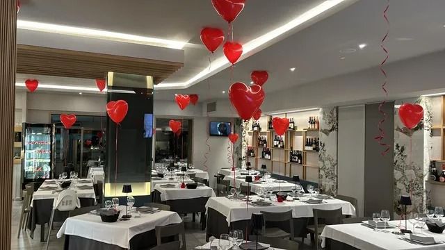L'interno di un ristorante è decorato con numerosi palloncini rossi a forma di cuore appesi al soffitto sopra i tavoli apparecchiati con tovaglie bianche.