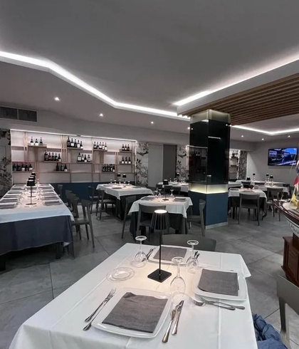 L'interno di un ristorante moderno e ben illuminato, con tavoli apparecchiati per la cena, tovaglie bianche, tovaglioli grigi e vino.