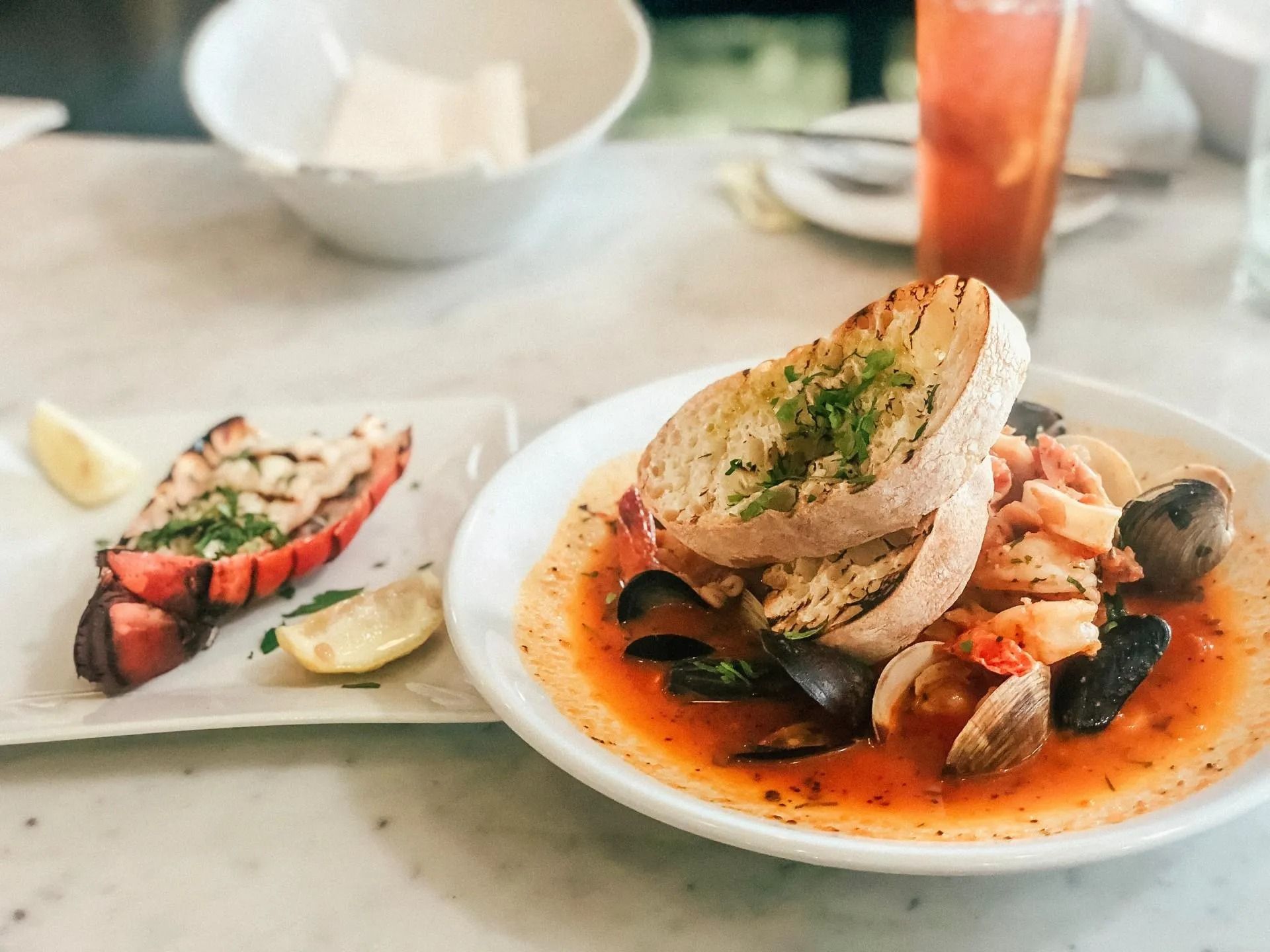 Una ciotola di zuppa di pesce con pane grigliato, accompagnata da una coda di aragosta grigliata su un piattino.