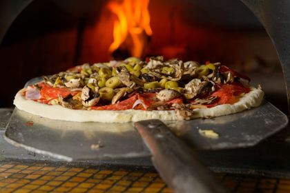 Una pizza condita con funghi e olive viene infornata in un forno a legna su una pala di metallo.