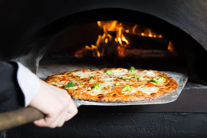 Una mano usa una pala di metallo per far scivolare una pizza condita con broccoli e formaggio fuso in un forno a legna con le fiamme.