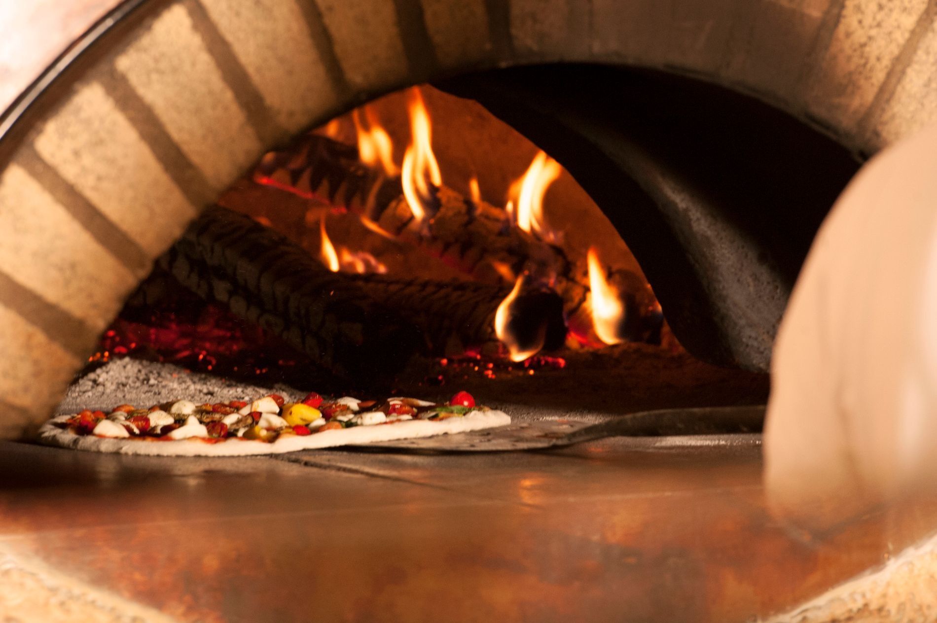 Una pizza cruda viene infornata in un forno a legna in mattoni, con fiamme vivaci sullo sfondo.