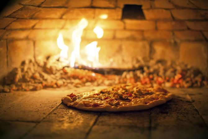 Una pizza che cuoce in un forno a legna in pietra, con fiamme e braci ardenti visibili sullo sfondo.