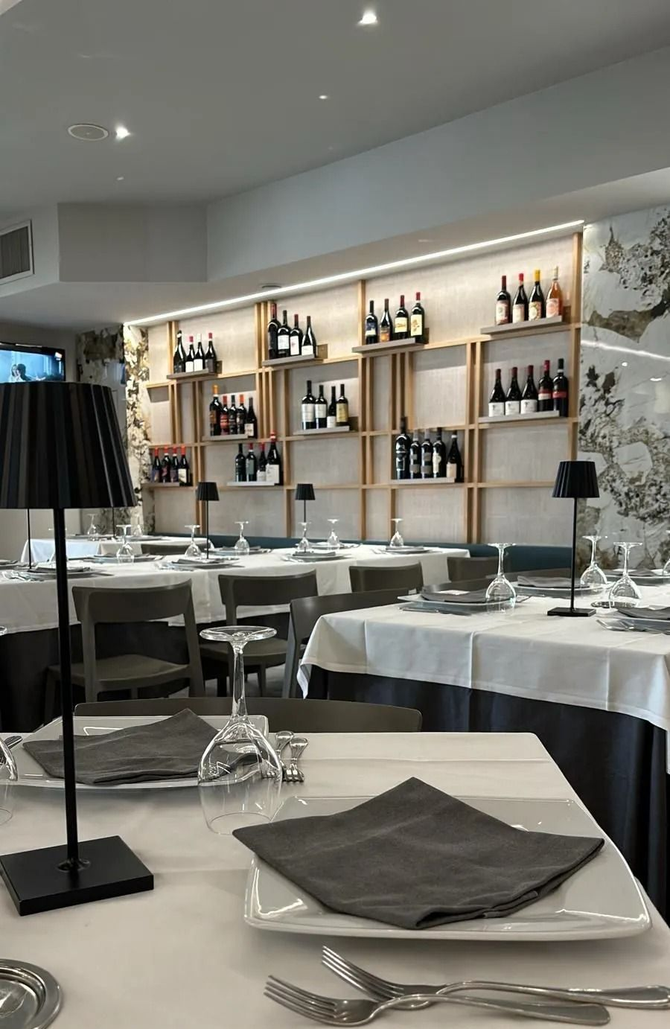 L'interno di un ristorante di lusso con tavoli apparecchiati con tovaglie bianche, lampade minimaliste e scaffali che espongono bottiglie di vino.