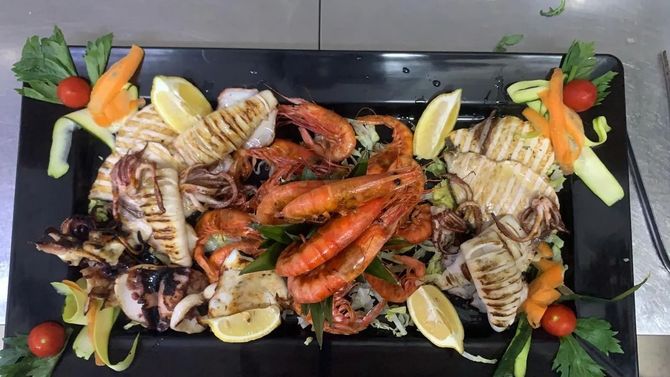 Un piatto rettangolare di gamberi e calamari grigliati, guarnito con spicchi di limone, sedano e pomodorini.