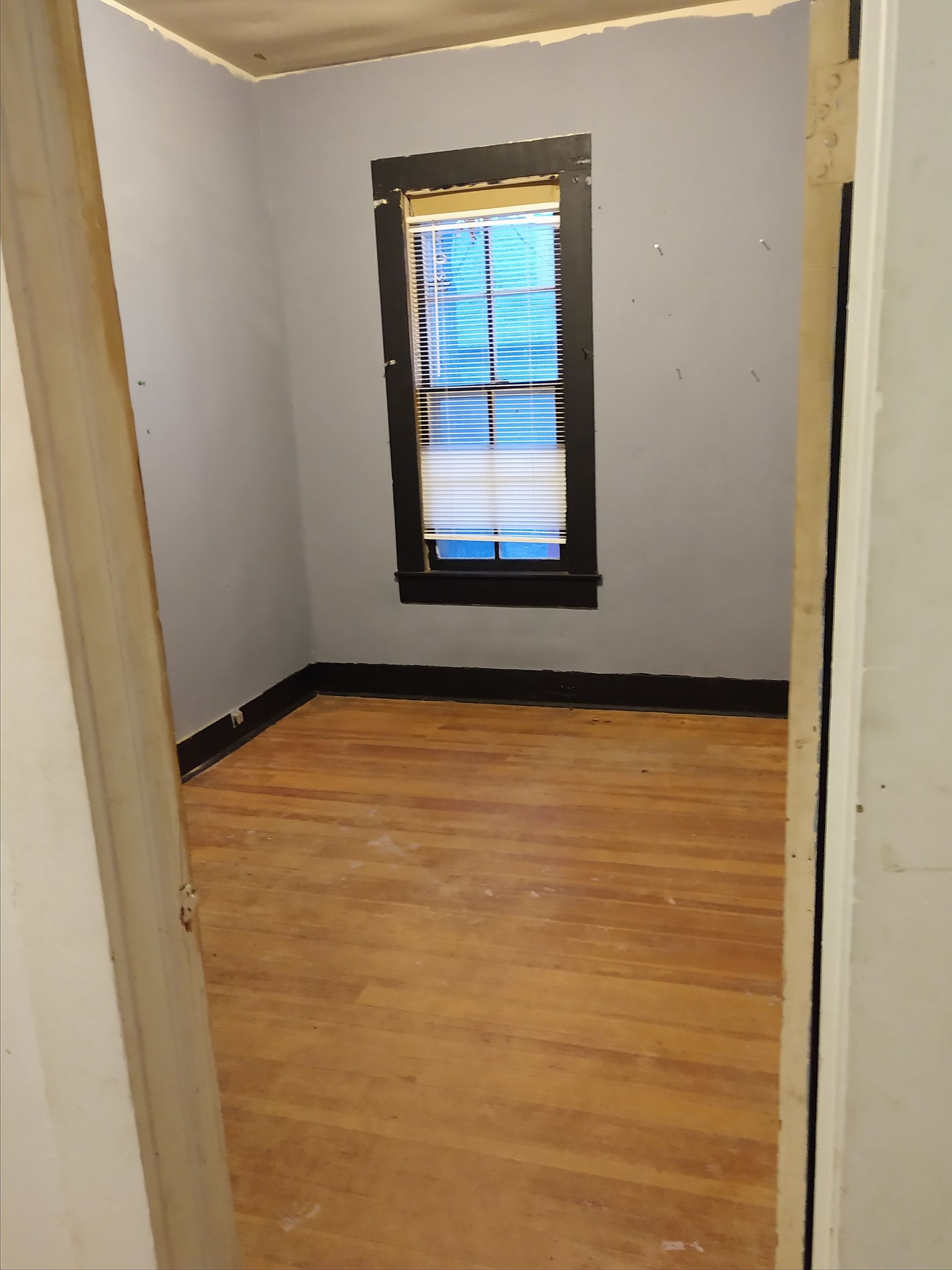 Small bedroom before remodel, with faded purple walls, black trim, sagging ceiling, and damaged wooden floors.