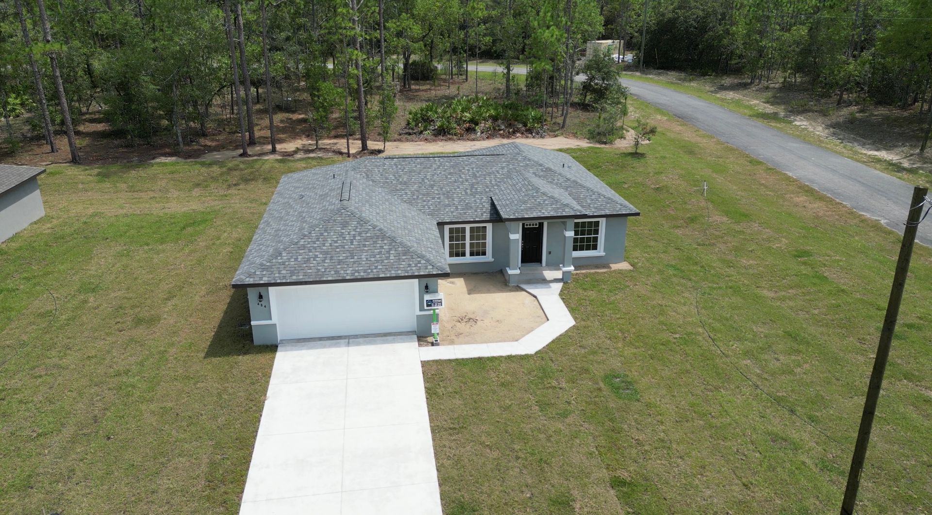Aerial view of a gray house with a white garage door, concrete driveway, and a grassy yard surrounded by trees.