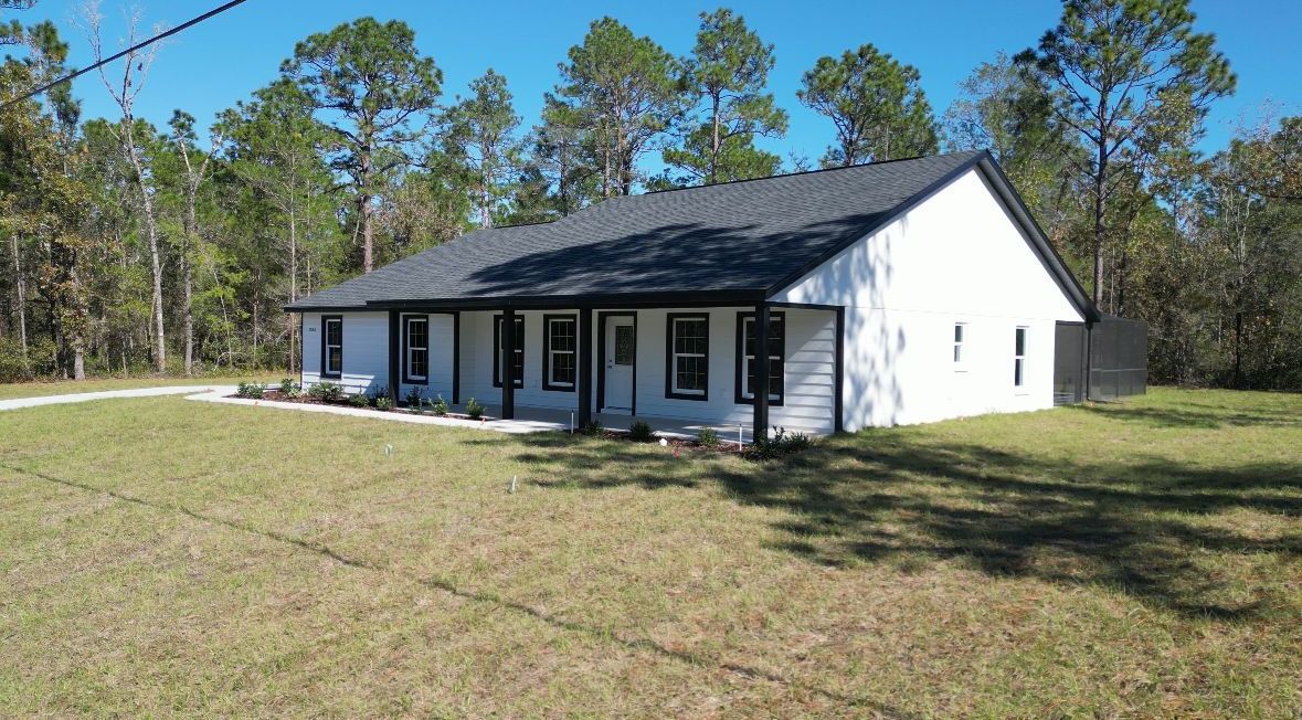 White house with black trim and roof in a grassy yard, surrounded by trees.