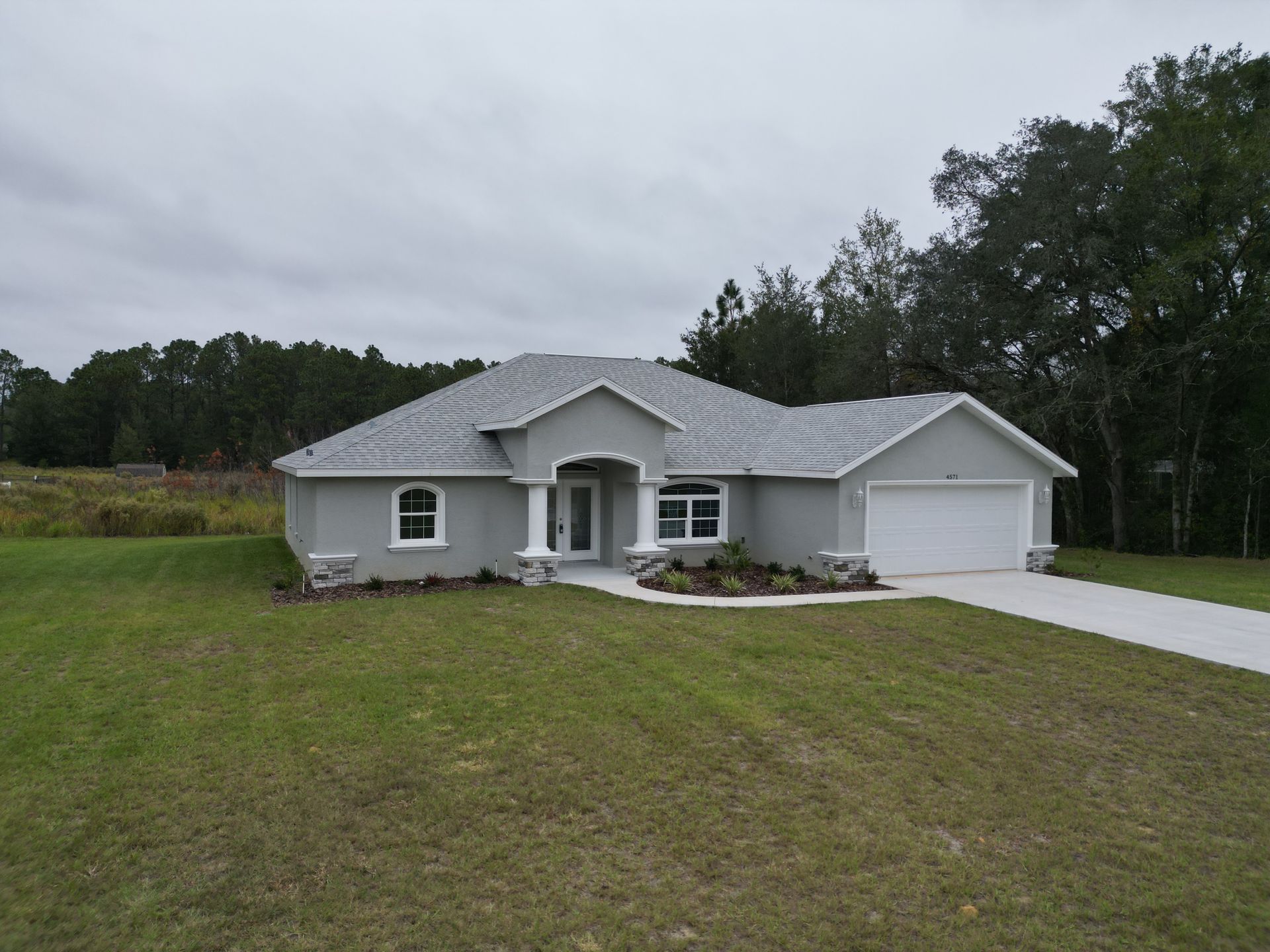 Gray stucco house with white trim, a driveway, and a lawn. Overcast sky and trees in the background.