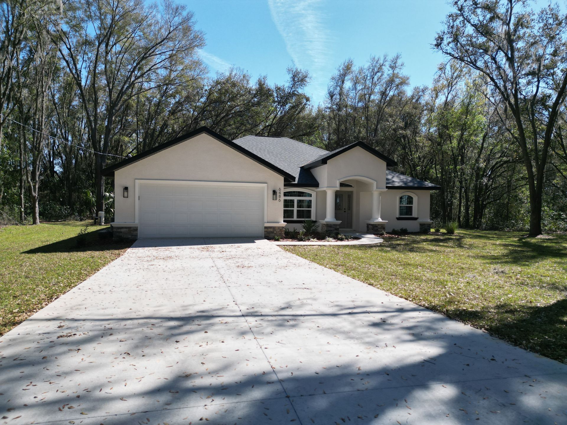 Beige house with white garage door and long concrete driveway, surrounded by trees.