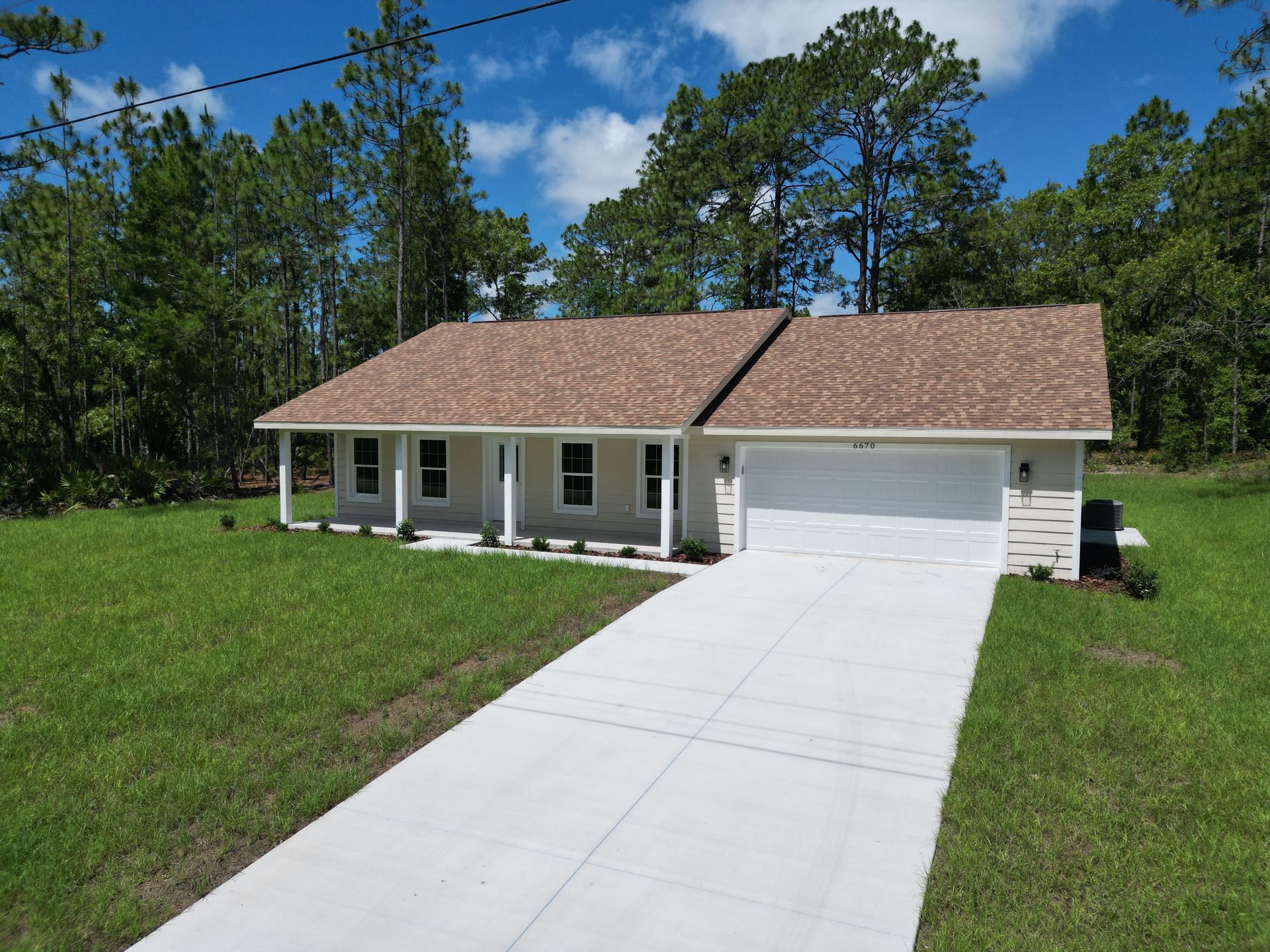 Light-colored house with a brown roof and white garage door, driveway. Green lawn, trees in background, blue sky.