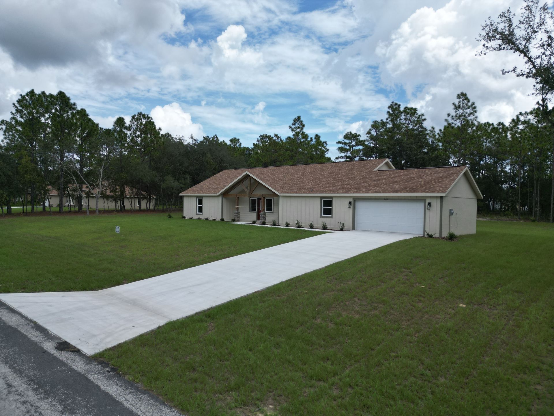 A single-story beige house with a long concrete driveway, set on a green lawn with a tree-lined backdrop and cloudy sky.