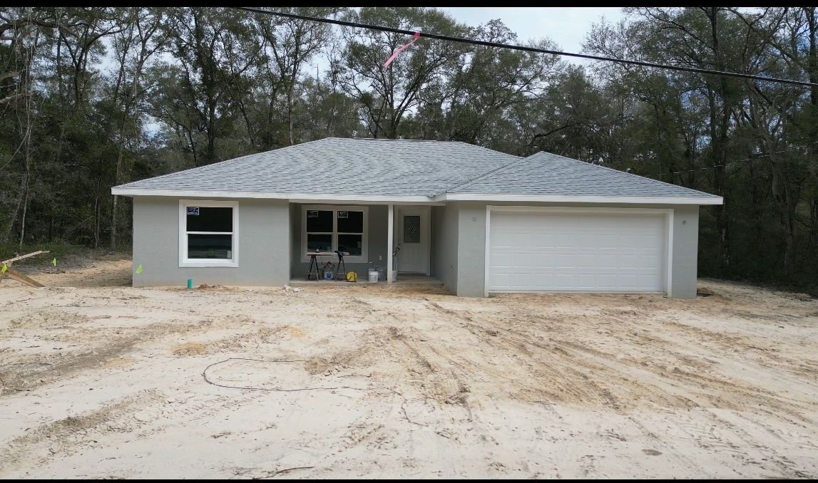 Gray, single-story house under construction. Sandy yard, white garage door, small windows.