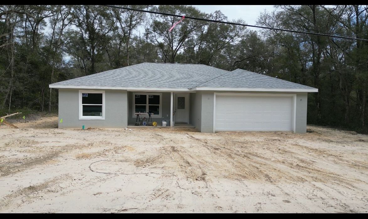 A light gray house with a two-car garage sits on a sandy lot, trees in the background.