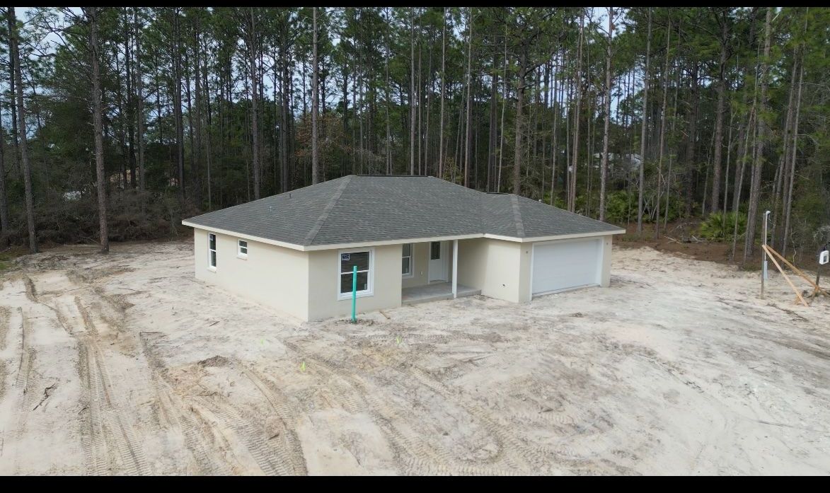 Beige house with gray roof, surrounded by sand, in a wooded area.