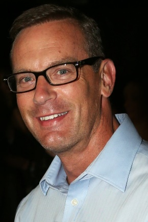 Smiling man wearing glasses and a light blue shirt against a dark background