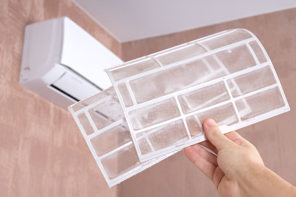 Person Holding Two Dirty Air Filters in Front of a Wall-mounted AC Unit — Frosty Solutions Air Conditioning In Mudgee, NSW