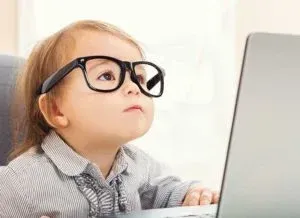 Child wearing large glasses looking up, using a laptop.