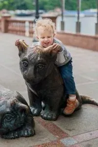 Child riding a bronze cat statue, smiling, outdoors.