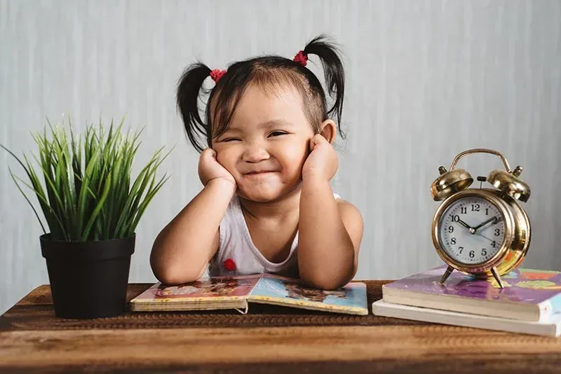 Smiling child resting head on hands at a desk with books, a plant, and a clock.
