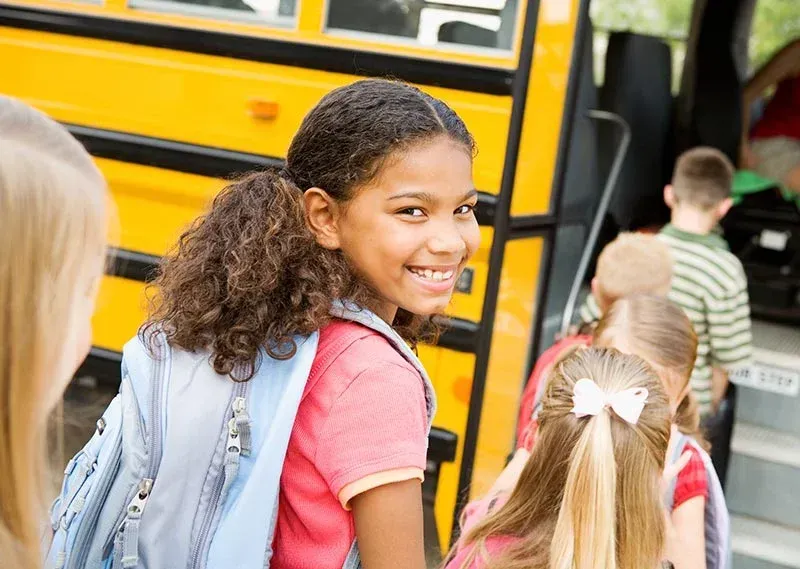 Girl with backpack smiles, boarding a yellow school bus with other children.