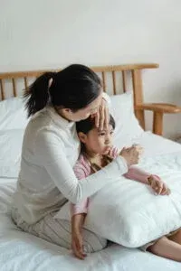 Woman checking child's forehead. Both sit on bed; child rests on a pillow.