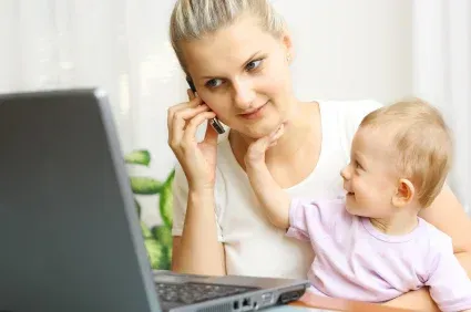 Woman holding baby while on phone, using laptop at a table. Baby reaches for woman's face; both have pleasant expressions.