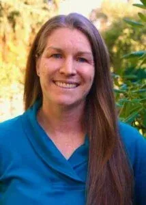 Woman with long brown hair smiles, wearing a blue shirt, outdoors.
