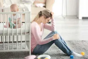 Woman sits on the floor, looking stressed, with a baby in a crib nearby.