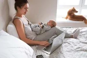 Woman working on laptop with baby on lap, dog on windowsill.