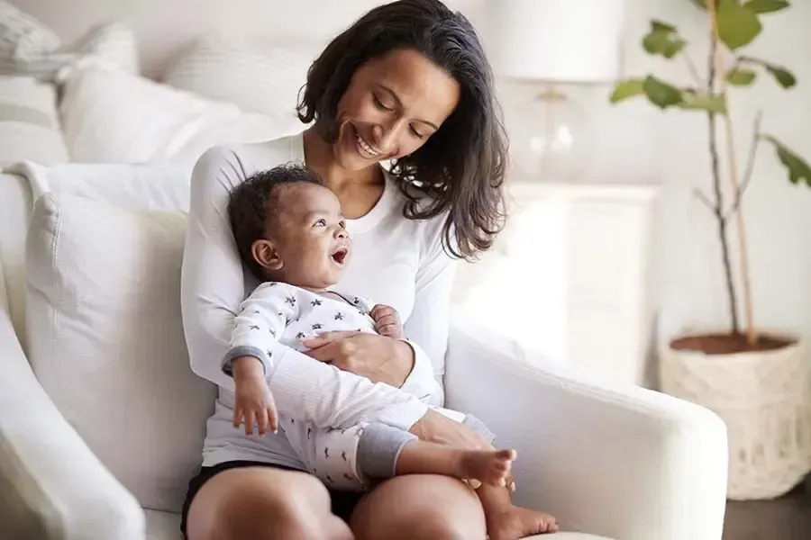 Woman holding and smiling at a baby in a white armchair.