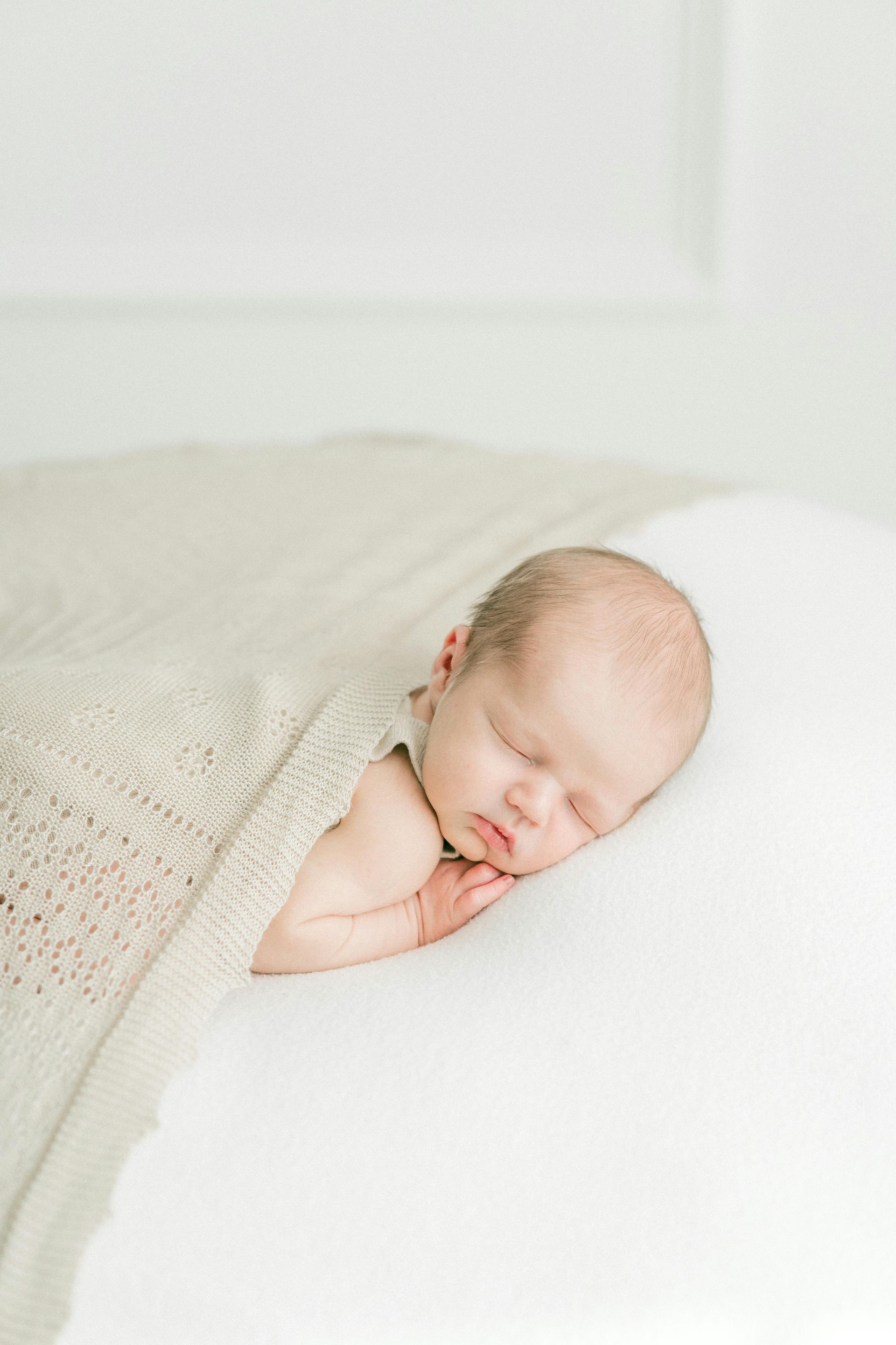 Newborn baby sleeping, swaddled in a light-colored blanket, on a white surface.