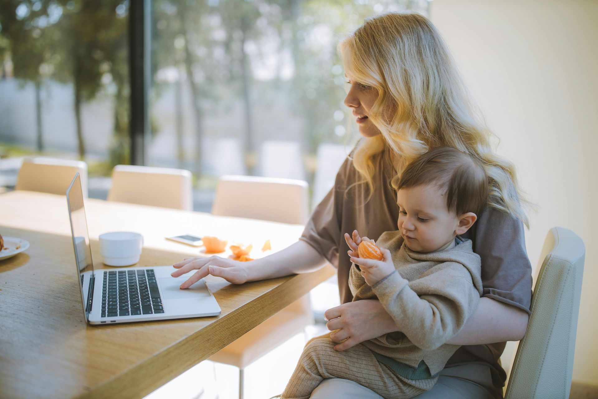 Woman working on laptop, holding a baby eating a snack at a table near a window.