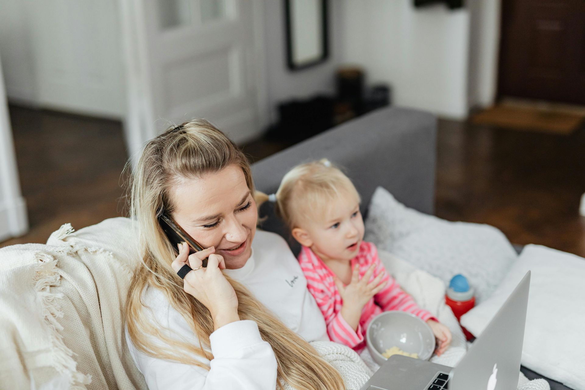 Woman on phone, working on laptop, with toddler sitting beside her on a sofa.