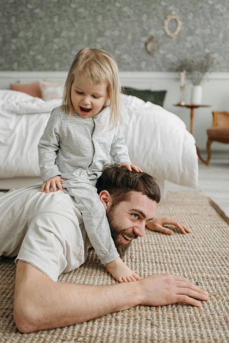 A person lies on a rug with a child sitting on their shoulders, both smiling in a bedroom.