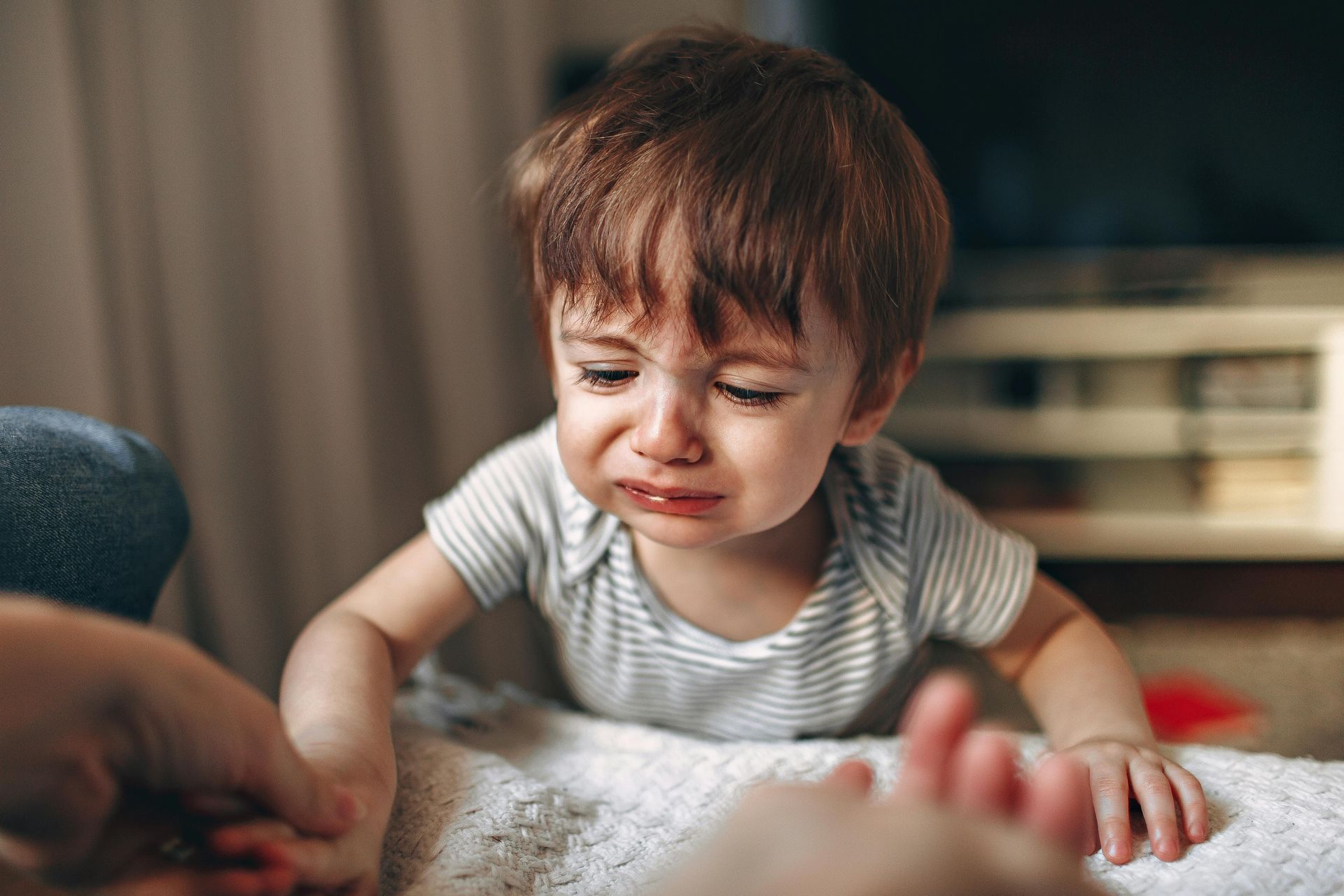 Crying toddler leans forward, reaching for a hand. Red hair, grey and white striped onesie. Indoors, blurry background.