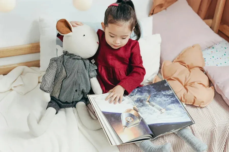 Girl reading a book with a stuffed animal on a bed with pillows.