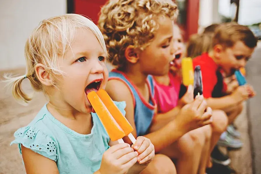 Children enjoying popsicles outdoors. Blonde girl licks orange popsicle, others eat theirs too.