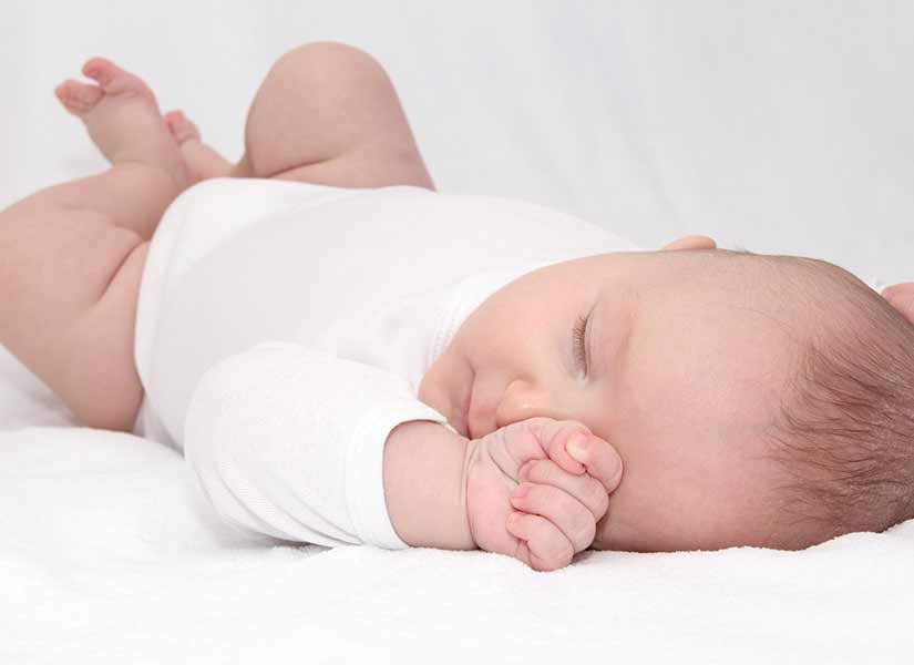 Baby sleeping on white sheets, wearing a white onesie, hand near face.