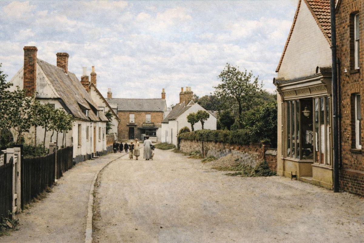 Quiet village street with cottages, a winding dirt road, and a few distant pedestrians under a cloudy sky