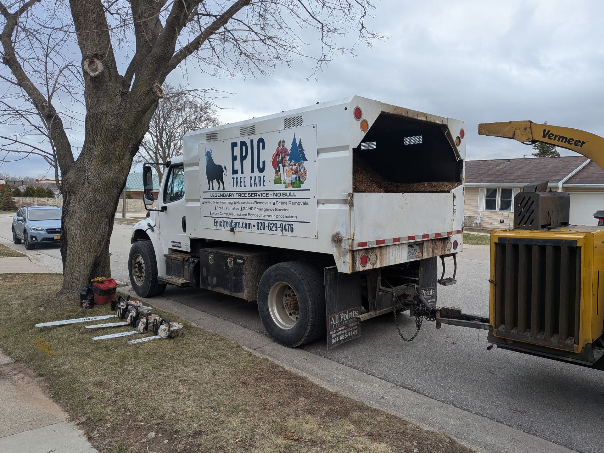 A white Epic Tree Service truck with a yellow wood chipper attached, parked on a residential street next to a tree.