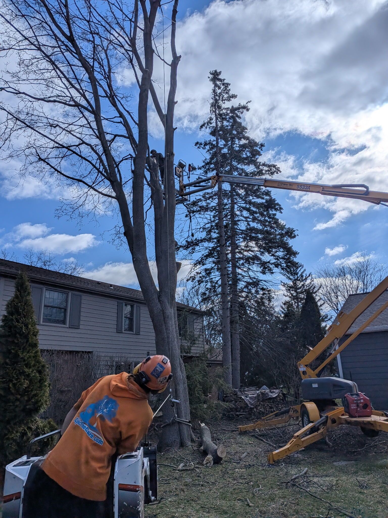 A worker in an orange hoodie and helmet operates a mechanical lift to trim a tree near a two-story house under a blue sky.